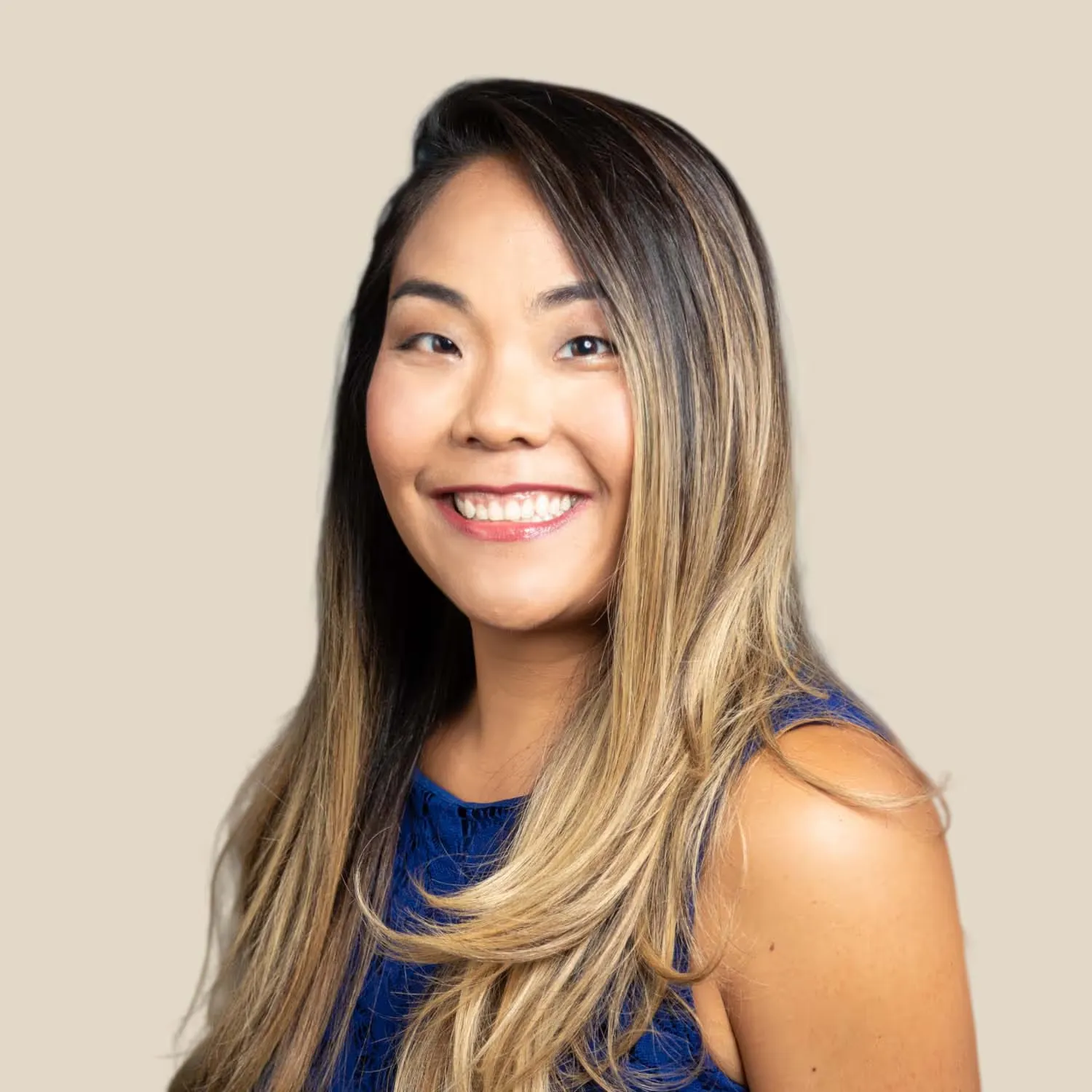 Smiling Asian woman with long, straight, highlighted hair wearing a royal blue sleeveless top, posed against a beige background.
